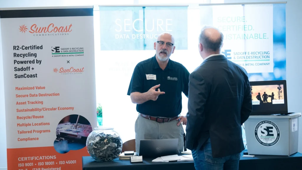 Two men conversing at a trade show booth with SunCoast and Sadoff E-Recycling banners.