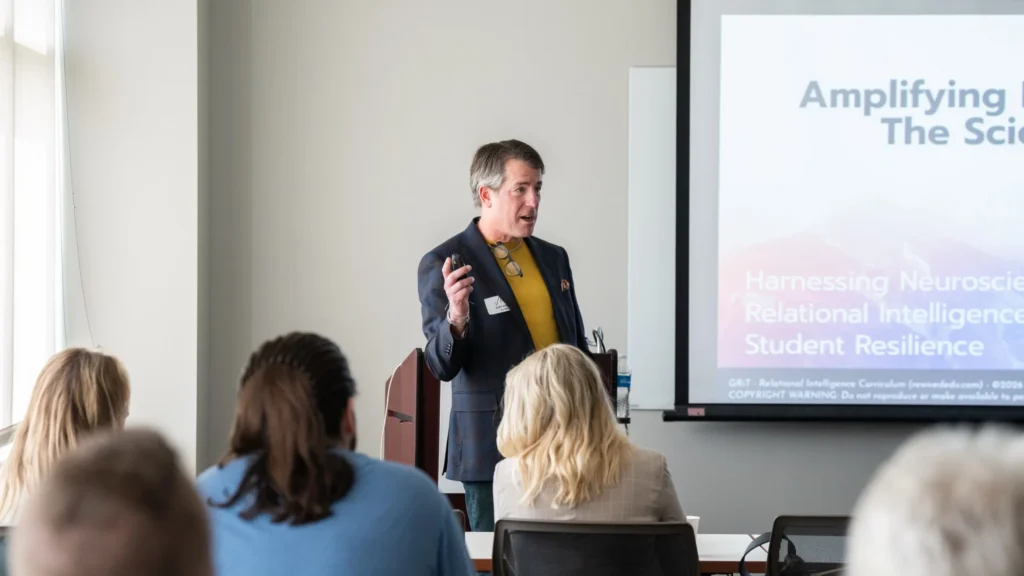 Man in blazer and yellow shirt presenting to seated audience, projection screen with text visible.