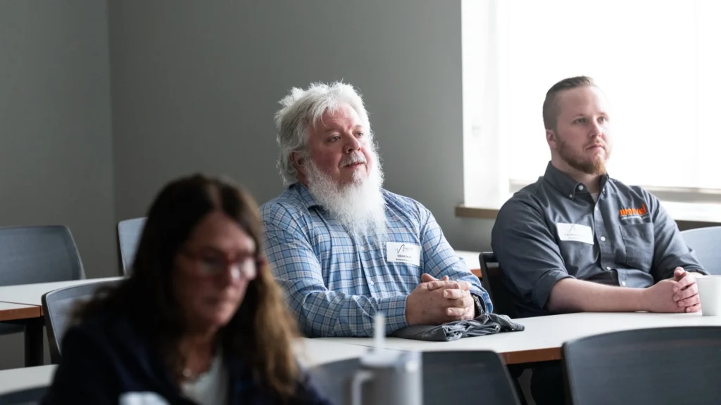 Three adults seated attentively in a classroom with name tags and casual attire.