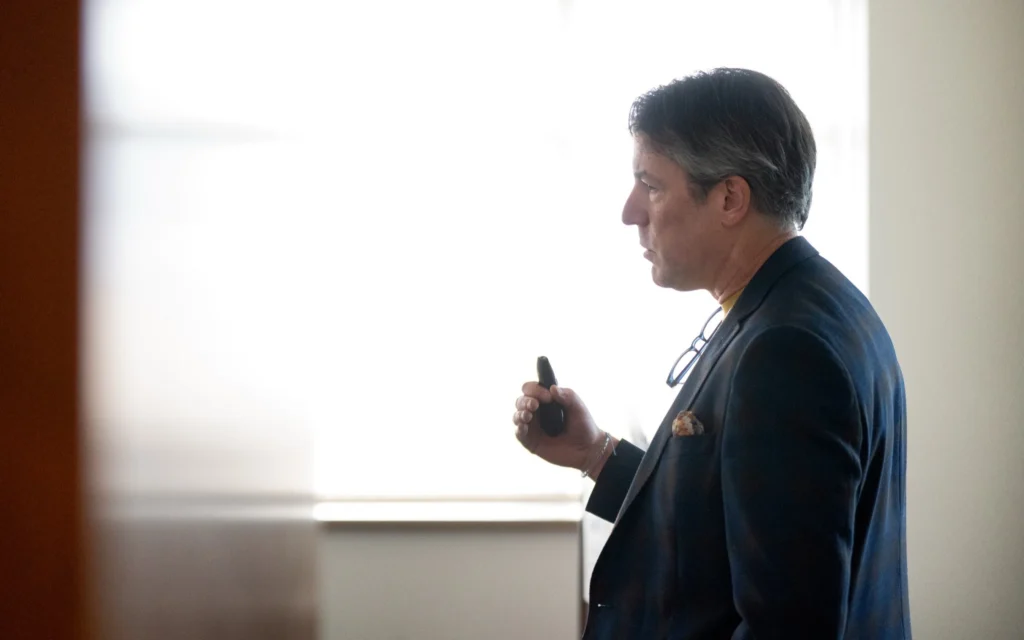 Middle-aged man in dark suit holding a remote, standing near a bright window.