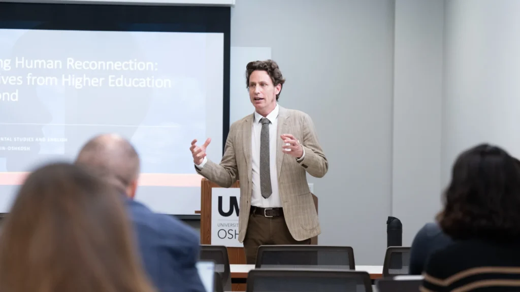 Man in beige suit jacket and tie giving presentation in classroom with projected slide and seated audience.