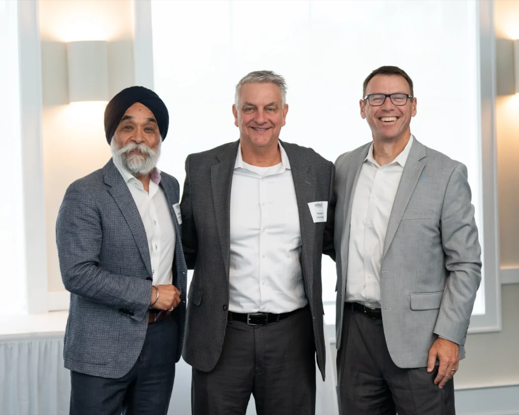 Three professionally dressed men smiling, standing indoors with neutral-colored walls and modern lighting.