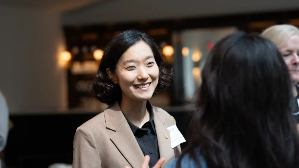 Professional Asian woman in beige blazer engaging in conversation at indoor event.