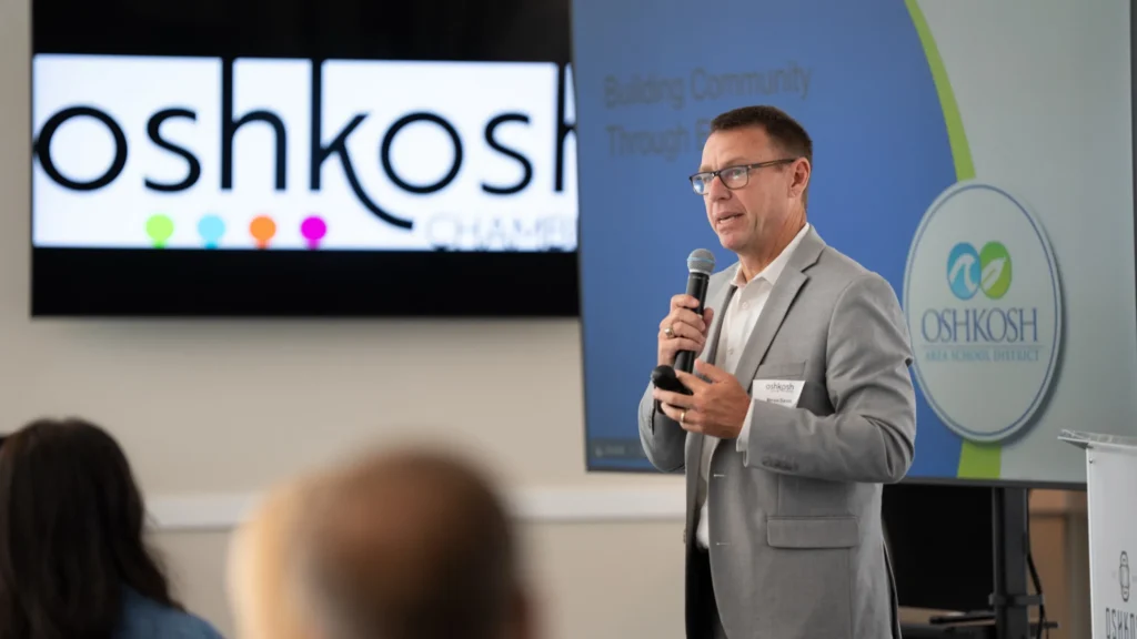 Professional man in gray suit speaking with microphone at Oshkosh Area School District presentation.
