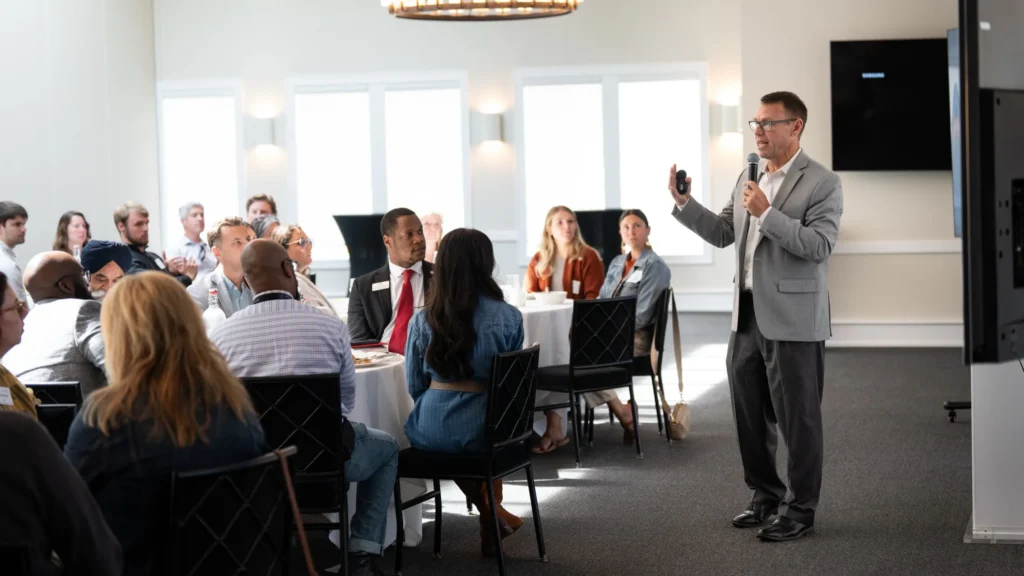 Professional speaker in gray suit presenting with microphone to diverse seated audience in conference room.