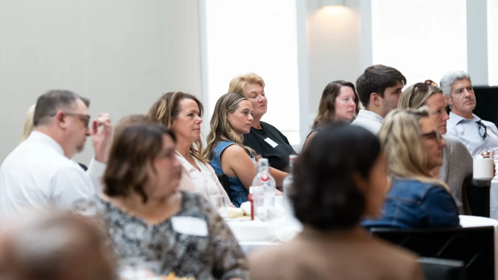 Group of diverse adults attentively seated at round tables in a bright conference room.