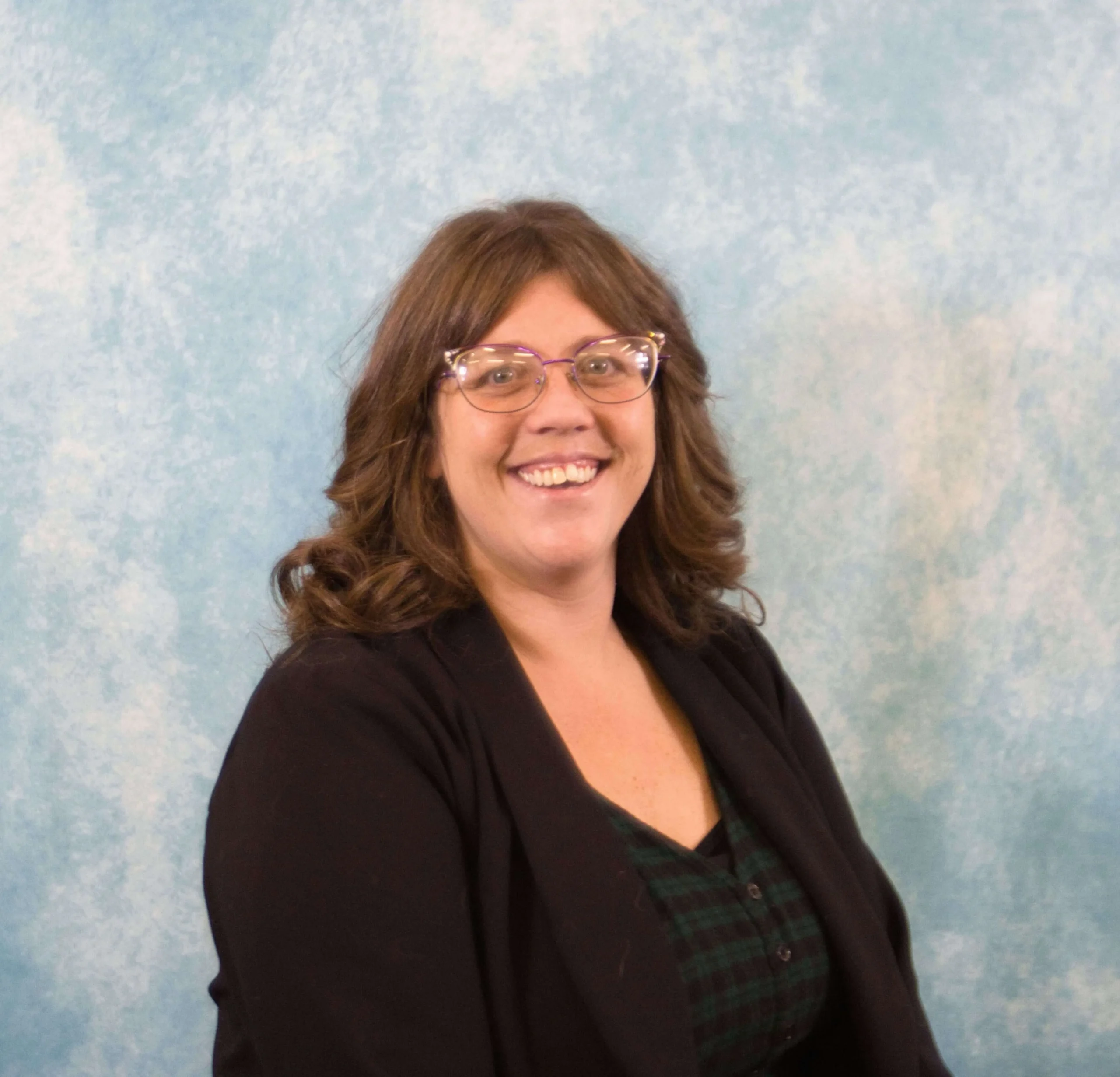 Professional portrait of a smiling woman with brown hair and glasses, wearing a black blazer.