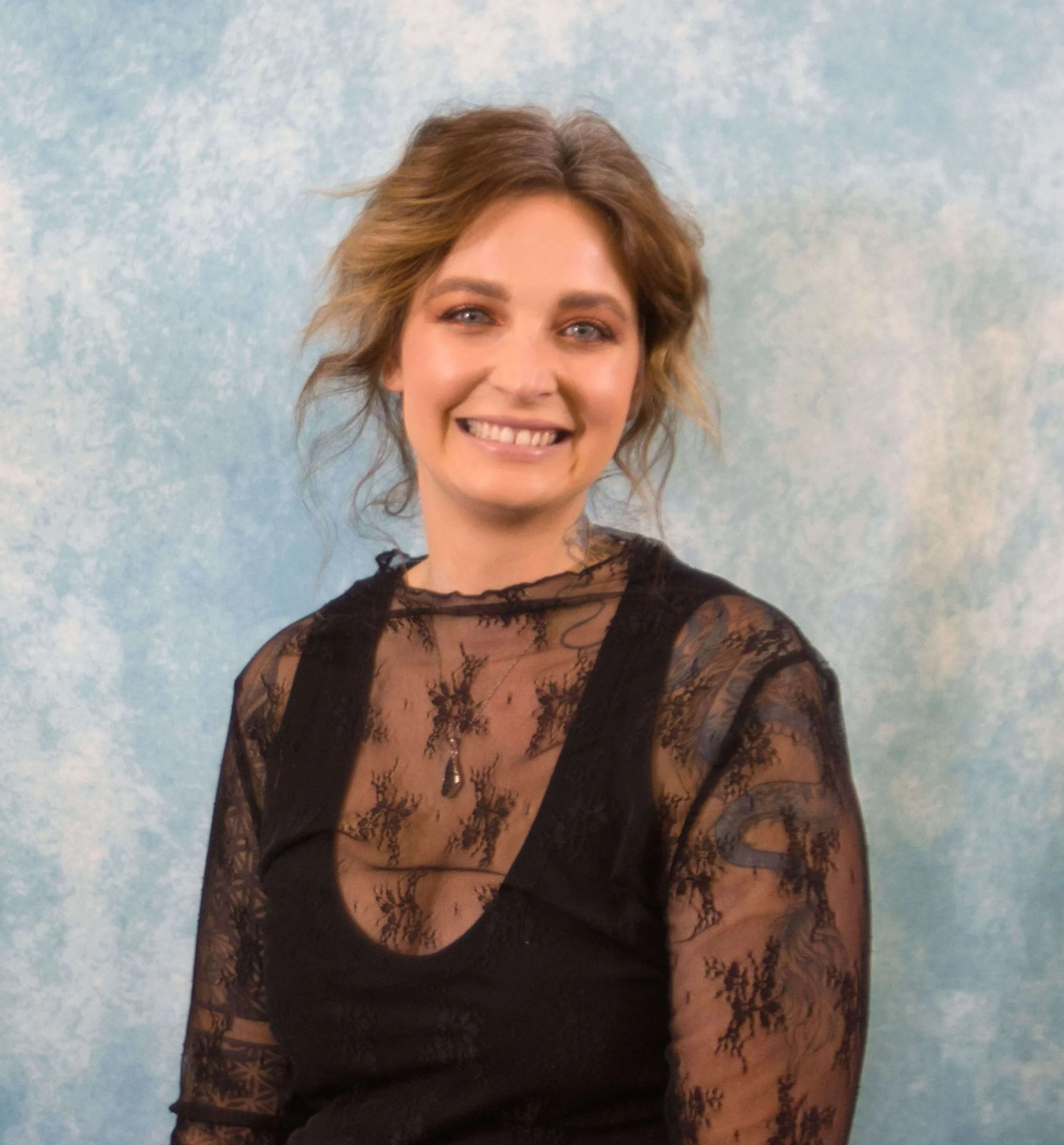 Portrait of a smiling woman with wavy brown hair wearing a black lace top against a blue textured background.
