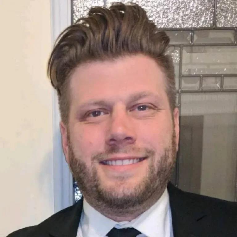 Portrait of a smiling man with styled brown hair and beard, wearing a black suit and white shirt.