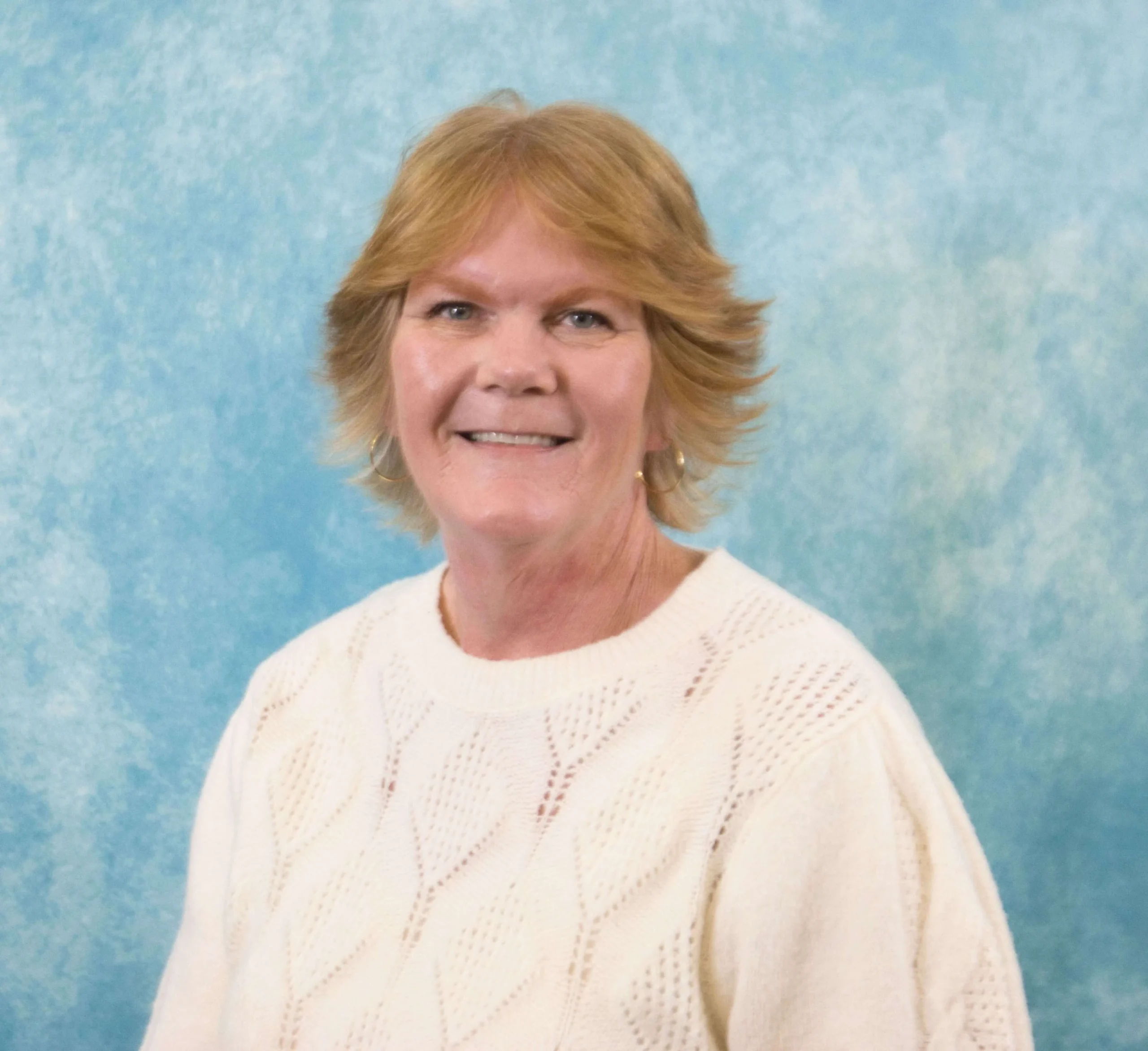 Portrait of a smiling middle-aged woman with short blonde hair, wearing a white knit sweater.