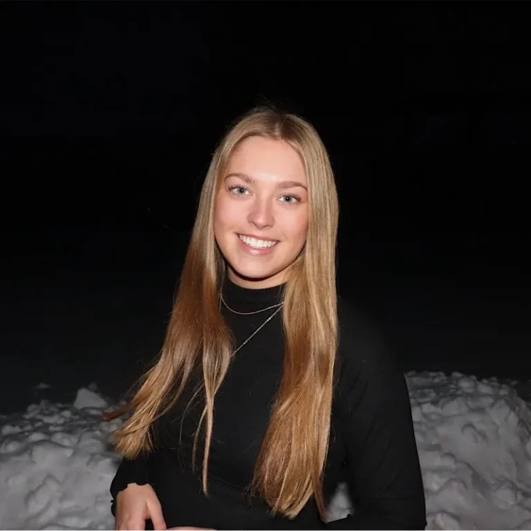 Young woman with long blonde hair wearing a black turtleneck, smiling against a snowy, dark background.