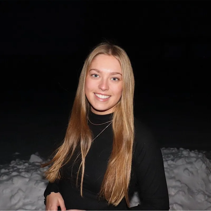 Young woman with long blonde hair wearing a black turtleneck, smiling against a snowy, dark background.