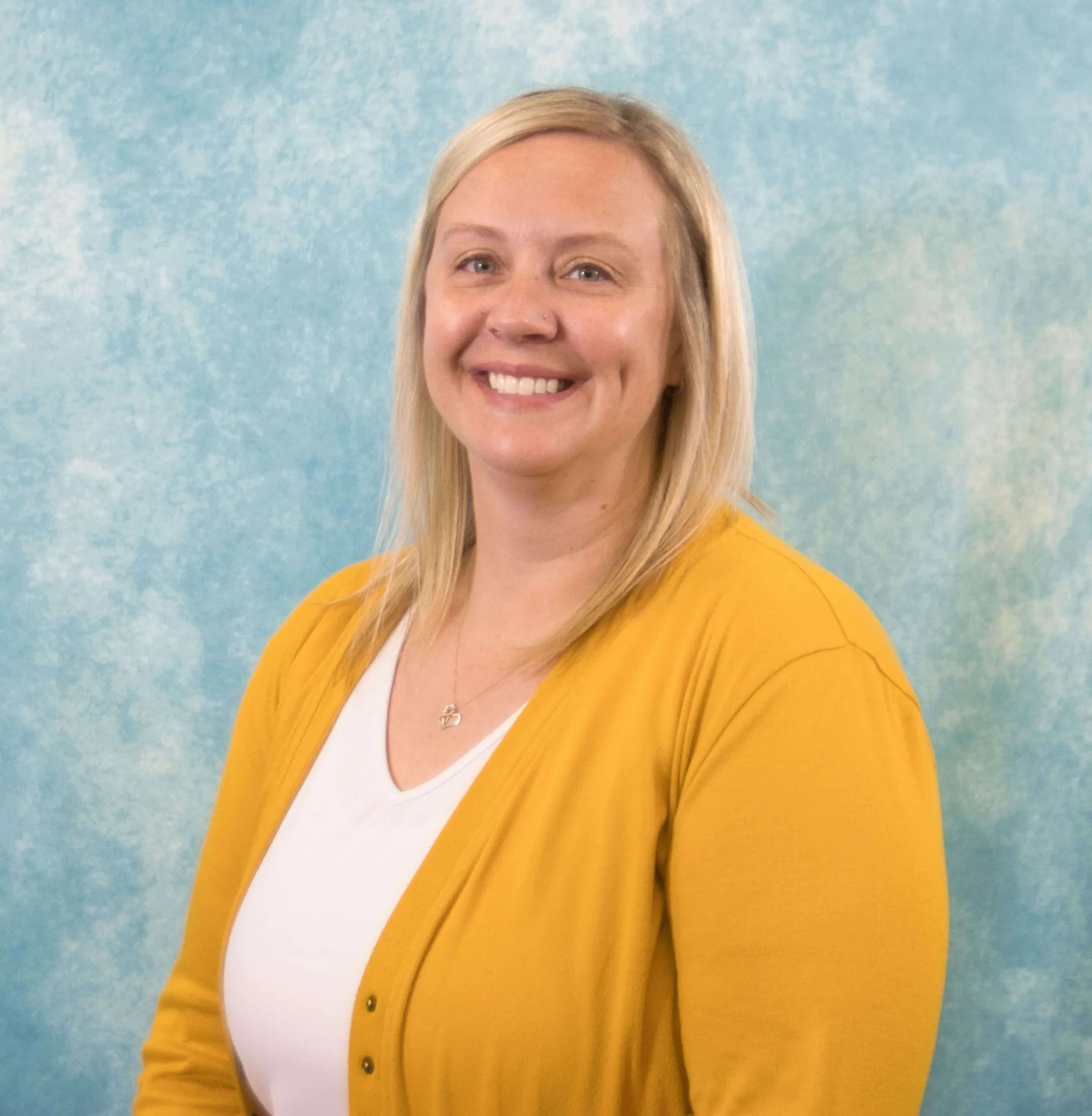 Portrait of a smiling woman with blonde hair, wearing a yellow cardigan and white top, blue textured background.