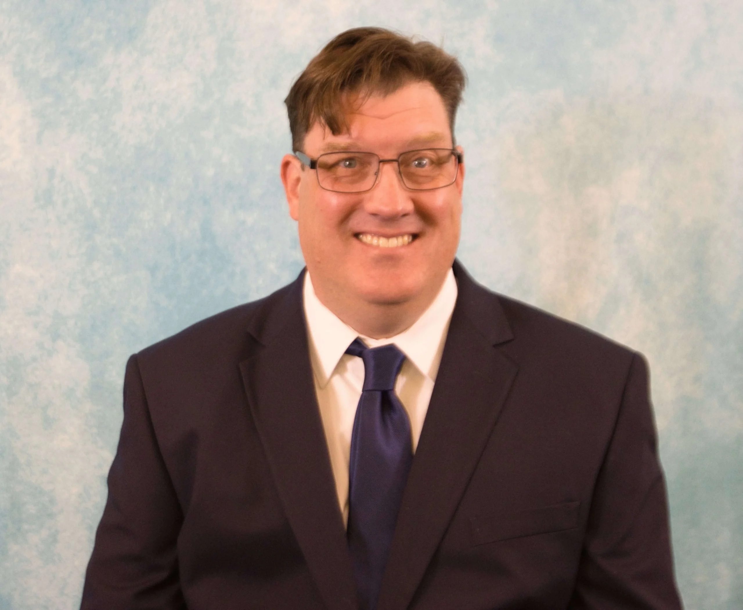 Portrait of a smiling man wearing glasses, a dark suit, white shirt, and blue tie against a light blue background.