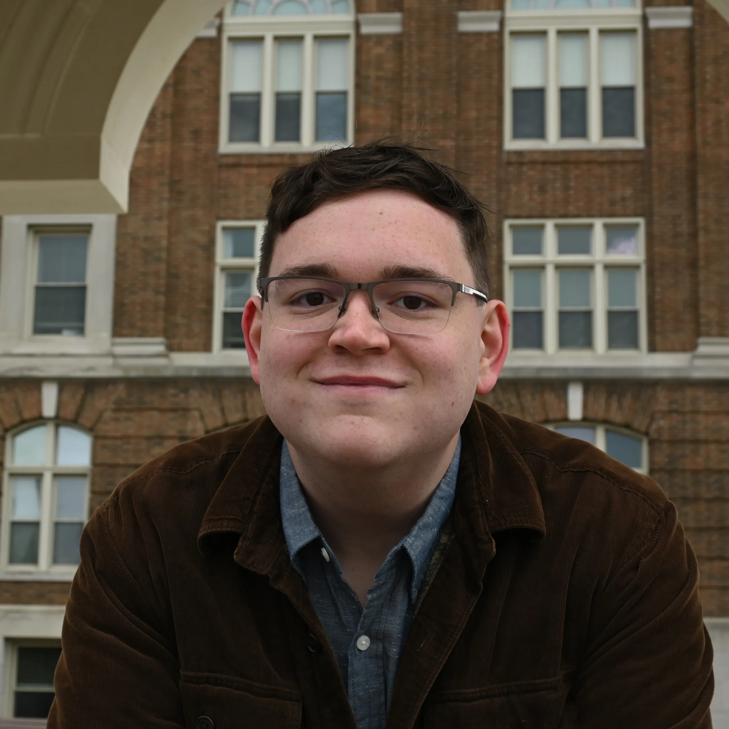 Portrait of a young man with glasses wearing a brown jacket in front of a brick building.