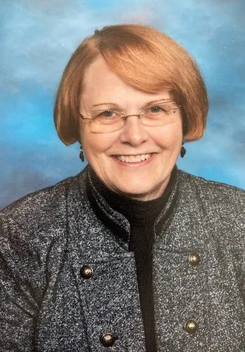 Portrait of a smiling middle-aged woman with short red hair, glasses, and a gray jacket.
