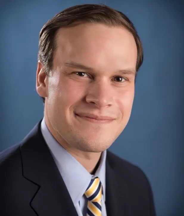 Professional headshot of a smiling man in a dark suit, light blue shirt, and striped yellow tie against a blue background.