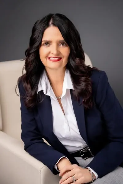 Professional portrait of a woman with dark hair, wearing a navy blazer and white blouse, seated on a beige chair.