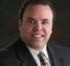Portrait of a man in a dark suit and patterned tie against a dark background.