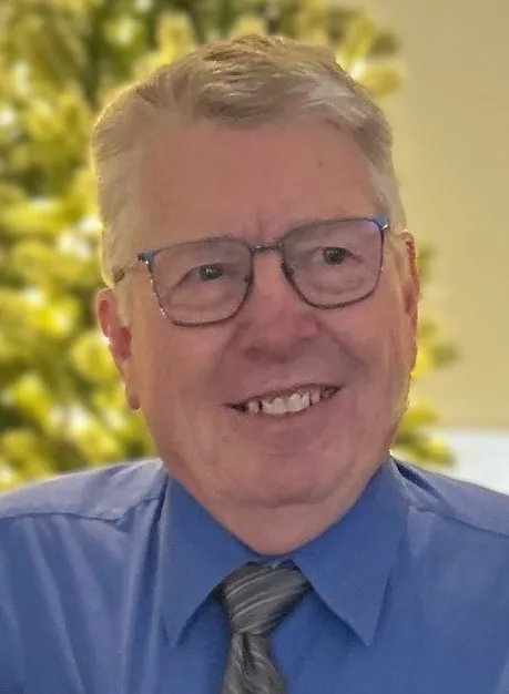 Portrait of an older man wearing glasses, a blue shirt, and a striped tie, with a blurred green background.