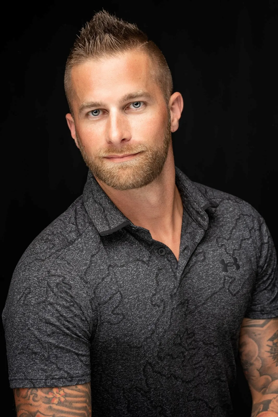 Portrait of a man with short spiked hair, beard, blue eyes, wearing a dark gray Under Armour shirt, with tattoos on his arms.